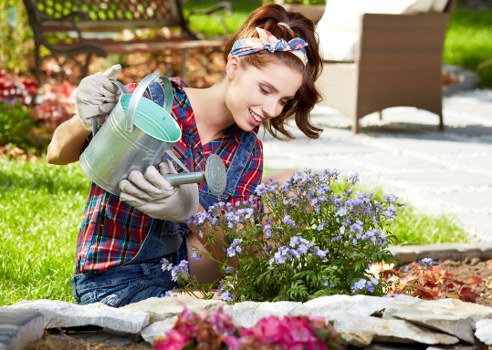 A young woman with light brown hair tied back and wearing a colorful plaid shirt, gloves, and a floral headband, is kneeling in a well-maintained garden in Bethnal Green, nurturing a flowering shrub with small purple blossoms. She holds a silver watering can in one hand, tilting it to water the plant, and a small gardening trowel in the other. The garden features a neatly edged flower bed bordered with natural stone, containing vibrant pink and purple flowers, alongside lush green grass and the flowering shrub. In the background, there is a wooden garden chair and a paved patio area, partially shaded by trees, under a clear sky, suggesting a bright, sunny day. The environment demonstrates a tidy outdoor space suitable for gardening and outdoor leisure, reflecting professional landscaping care that a company like Landscapers Bethnal Green might provide for local homeowners seeking garden maintenance and enhancement.