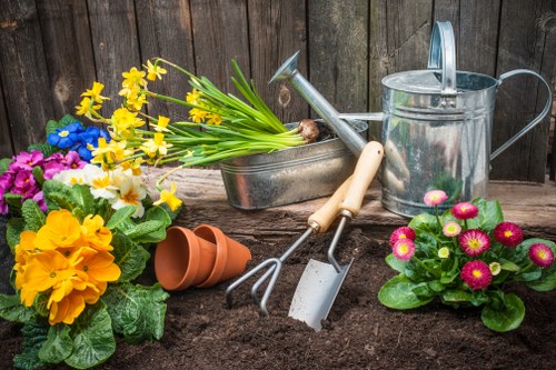 A freshly turned garden bed with dark, rich soil is bordered by a weathered wooden fence in the background. To the left, vibrant yellow and purple flowering plants are displayed, while on the right, bright pink and white primulas add colour to the scene. Gardening tools including a small hand rake, trowel, and watering can are arranged on the soil, alongside empty terracotta plant pots and a metal container holding a bunch of yellow daffodils. The outdoor space appears well-maintained, ready for planting or future landscaping work, reflecting typical garden care activities in Bethnal Green, with natural lighting suggesting a clear day. This setup exemplifies a typical gardening scene suitable for a professional landscaping or garden maintenance service to enhance outdoor spaces around London E2 postcode area, as part of services provided by Landscapers Bethnal Green.
