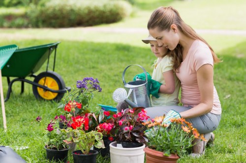 The image depicts a mother and her young daughter engaging in gardening activities in a lush, well-maintained backyard. They are kneeling on a vibrant green lawn with neatly trimmed grass, surrounded by a variety of potted flowering plants and shrubs that display bright red, purple, and pink blossoms, adding colourful accents to the outdoor space. In the background, a garden wheelbarrow with yellow wheels is positioned on the grass, indicating active outdoor maintenance. The garden area is bordered by natural vegetation and a blurred backdrop of trees and hedges, creating a peaceful, private environment. The mother appears to be guiding her daughter in watering or tending the plants, highlighting a moment of outdoor activity and nature connection. The scene is illuminated by natural daylight, suggesting a clear, mild weather day suitable for gardening and lawn care tasks, consistent with professional gardening services like those offered by Landscapers Bethnal Green. This setting exemplifies a productive and aesthetically pleasing garden layout, combining lawn care, planting, and outdoor nurturing in a typical residential outdoor space near Bethnal Green, London.