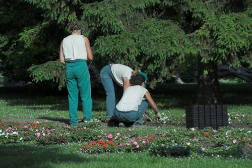 Two individuals are tending to a flower bed in a lush backyard garden, with a large tree providing shade overhead. One person is crouching while planting or weeding among multicoloured flowers, and another is kneeling nearby, engaged in similar gardening tasks. The garden features a well-maintained grassy lawn, bordered by a flower bed filled with vibrant blooms and delicate foliage. In the background, a dense arrangement of mature trees with deep green leaves offers natural privacy and a serene outdoor environment. The scene is set on a clear day, with natural sunlight highlighting the rich colours of the flowers and the textures of the lawn and tree bark, emphasizing a peaceful, well-cared-for landscaped space consistent with professional gardening and landscaping services in Bethnal Green.