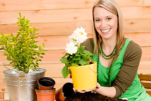The image shows a smiling woman with blonde hair, sitting outdoors in a garden area with a wooden fence in the background. She is holding a yellow flower pot containing a blooming white azalea or garden shrub. On her right, there are various gardening pots, including a black plastic pot and an empty terracotta pot, placed on a surface along with a small silver metal container holding a healthy green shrub, possibly a small hedge or ornamental plant. The setting appears to be a well-maintained garden or backyard space, with natural daylight illuminating the scene, and the environment suggests active gardening or plant care. The overall scene highlights elements such as potted plants, flower beds, and garden containers indicative of outdoor landscaping or garden maintenance services by Landscapers Bethnal Green, aligning naturally with gardening and outdoor care in the local area.
