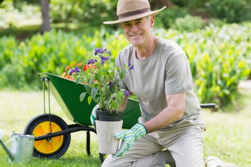 A smiling elderly man wearing a light grey t-shirt, wide-brimmed straw hat, and gardening gloves kneels on a well-maintained lawn in a garden in Bethnal Green, holding a potted plant with vibrant purple, red, and pink flowers. Behind him, there is a bright green wheelbarrow with large yellow and black wheels, partially visible, placed on a grassy area. The garden features a neatly trimmed hedge or shrubbery in the background, along with tall trees and lush greenery that suggest a serene outdoor space, possibly a backyard or communal garden. The scene is illuminated by natural daylight, indicating clear weather. The setting reflects a typical landscaped outdoor area suitable for gardening and lawn care services offered by Landscapers Bethnal Green, highlighting the importance of maintaining healthy turf, flower beds, and vibrant plantings in residential or communal outdoor spaces in the local area.