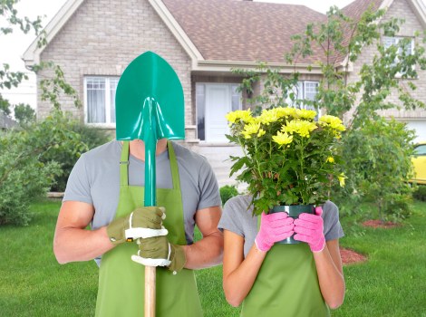 A gardener and a woman are standing in a well-maintained front garden in Bethnal Green during daylight, both wearing green aprons and gardening gloves; the gardener is holding a large green shovel with a shiny metal blade, while the woman is holding a pink watering can filled with bright yellow flowering plants, obscuring her face. The garden features a neatly trimmed lawn, a variety of green shrubs, and trees with fresh spring foliage. In the background, there is a light-colored brick house with white-framed windows and a sloped roof, surrounded by small flower beds and landscape edging. The environment appears bright and overcast, suggesting mild weather suitable for outdoor gardening tasks. This scene reflects typical gardening and landscaping activities, aligning with services offered by Landscapers Bethnal Green, with a focus on planting, lawn care, and outdoor maintenance in the local area.
