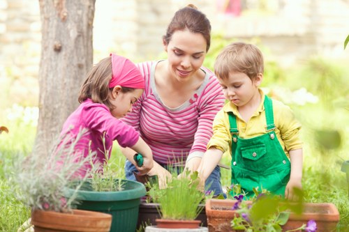A woman with dark hair pulled back, dressed in a pink and white striped top, is outdoors in a garden with two young children, one girl wearing a pink headscarf and a purple top, and one boy in a yellow top with green overalls. They are engaged in gardening activities, tending to various potted plants and flower beds positioned on a wooden garden table. The garden features lush greenery with grass, a large tree trunk visible in the background, and a mixture of flowering plants and foliage arranged along the border. The scene is illuminated by natural sunlight, creating a warm and inviting outdoor environment typical of a well-maintained garden space in Bethnal Green, which borders East London, and showcases a clean, organized area suitable for outdoor gardening and planting activities, reflecting professional gardening and landscaping practices by Landscapers Bethnal Green.