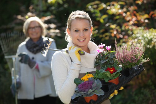 A woman with blonde hair, wearing a cream sweater and yellow gardening gloves, is holding a black tray filled with colorful flowering plants including purple, yellow, and red blooms, in a well-maintained garden setting. Behind her, an older woman with short blonde hair, glasses, a light jacket, and a scarf is slightly out of focus, holding garden tools such as a small rake and pruners. The garden features a lush, green landscape with various shrubs, trees with autumn-colored leaves, and a neatly trimmed lawn, suggesting an outdoor space well cared for by professional landscapers like Landscapers Bethnal Green. The scene is illuminated by natural daylight, highlighting the vibrant plant colors and healthy foliage, indicative of outdoor garden maintenance and planting services in Bethnal Green's residential areas, possibly within the postcode district surrounding E2, contributing to a welcoming and thriving garden environment.